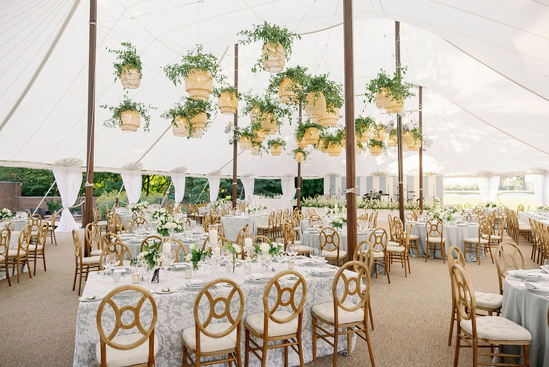 A White Tent, Many Tables With White Tablecloth, Wood Chairs, And Rattan Lanterns Wedding Decor Above