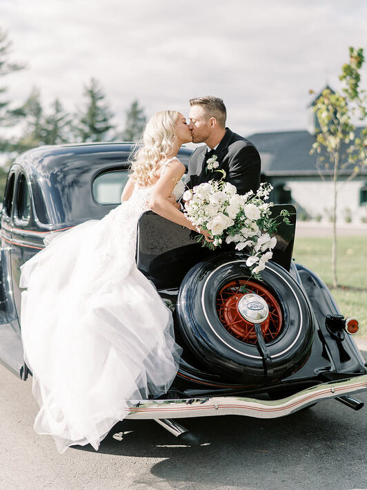 The Bride And Groom Kiss While On Top Of The Back Trunk Of A Black Classic Car