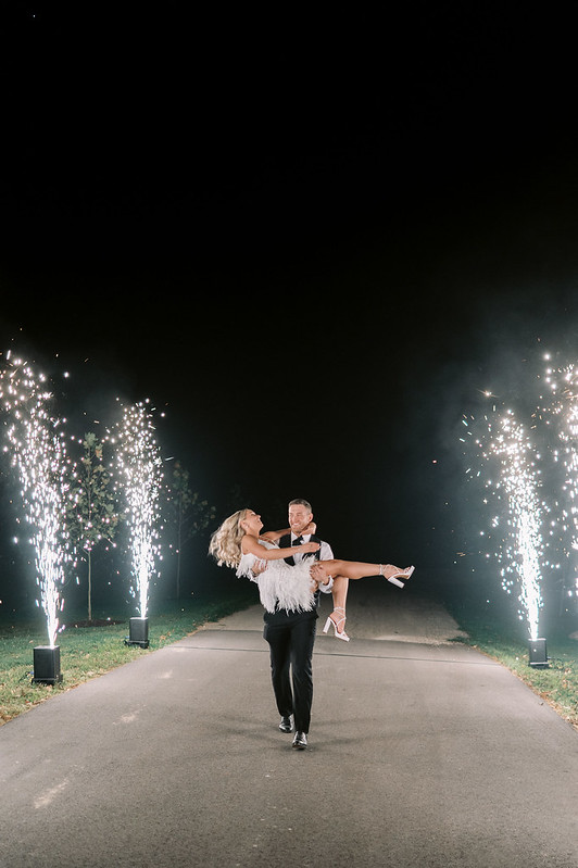 A Joyful Groom Carrying His Bride, With A Cold Spark Machine Lighting Up The Scene At The Clubhouse Wedding