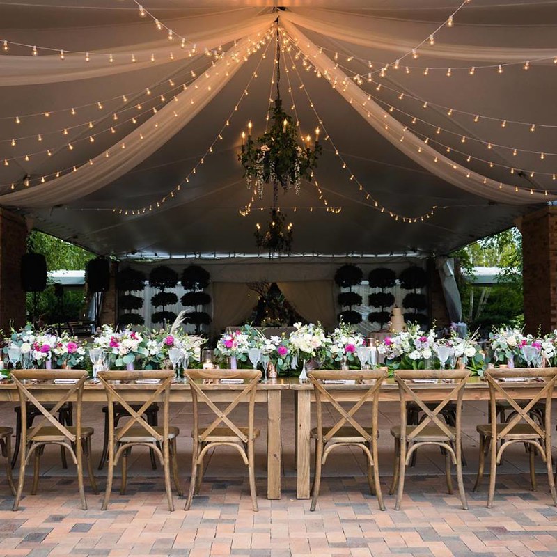 Wedding Tent With Hanging String Lights, Long Tables With Floral Decor And Wooden Chairs