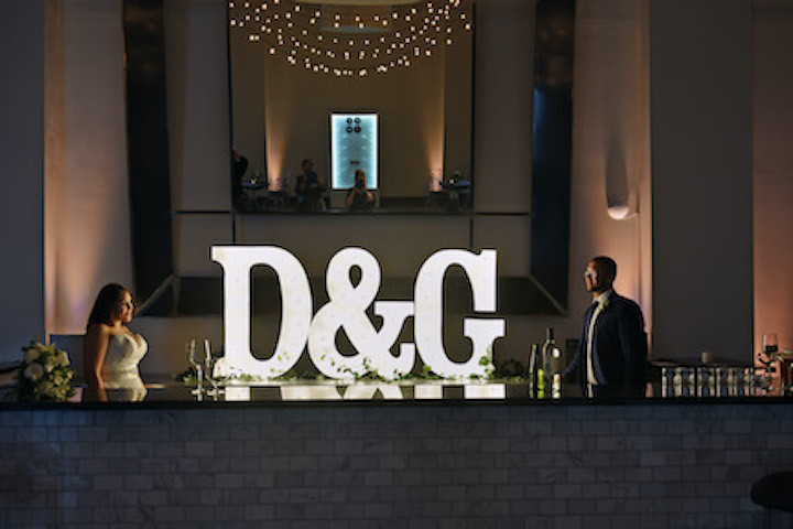 Bride And Groom Taking Picture With Marquee Letters Spell Out Their Initials As Their Backdrop