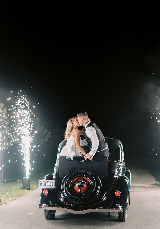 The Bride And Groom Sharing A Kiss While On Top Of A Black Vintage Classic Car