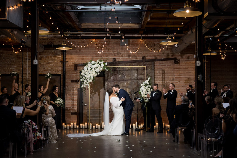 Bride And Groom Shared A Kiss Under Draping String Lights, Guests Happily Watching Them