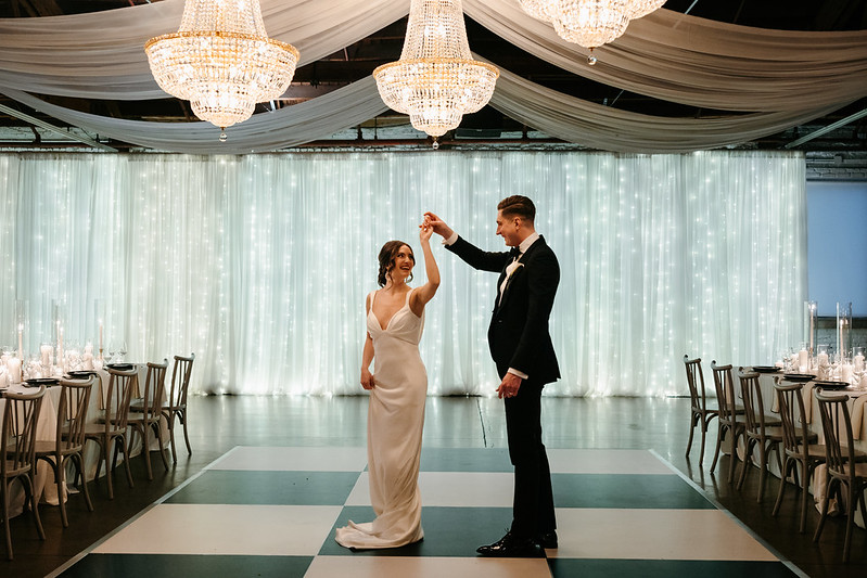 Bride And Groom Dancing Under Elegant Chandeliers, Black And White Checkered Dancefloor, And Twinkle Lights Backdrop