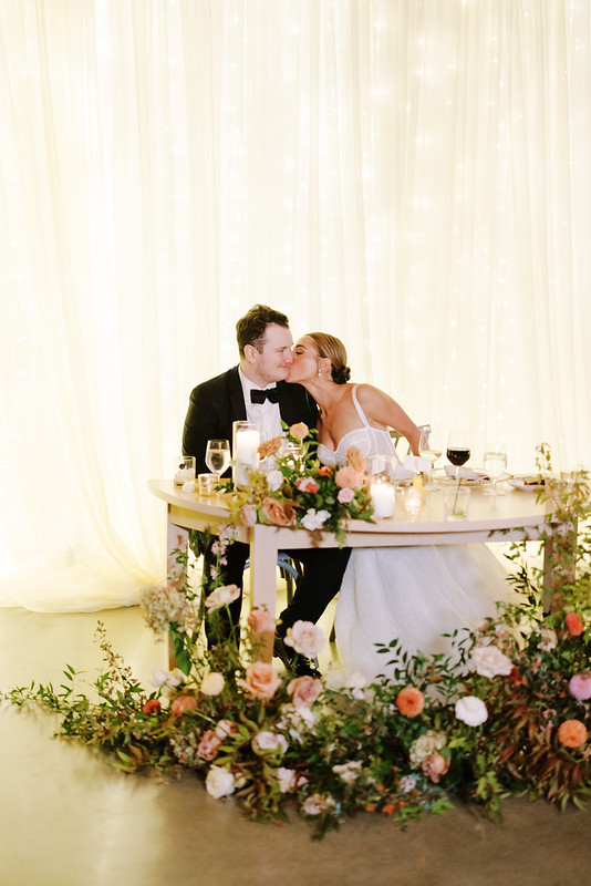 Hannah And Declan Sitting At The Head Table With Drape Twinkle Lights As Their Backdrop